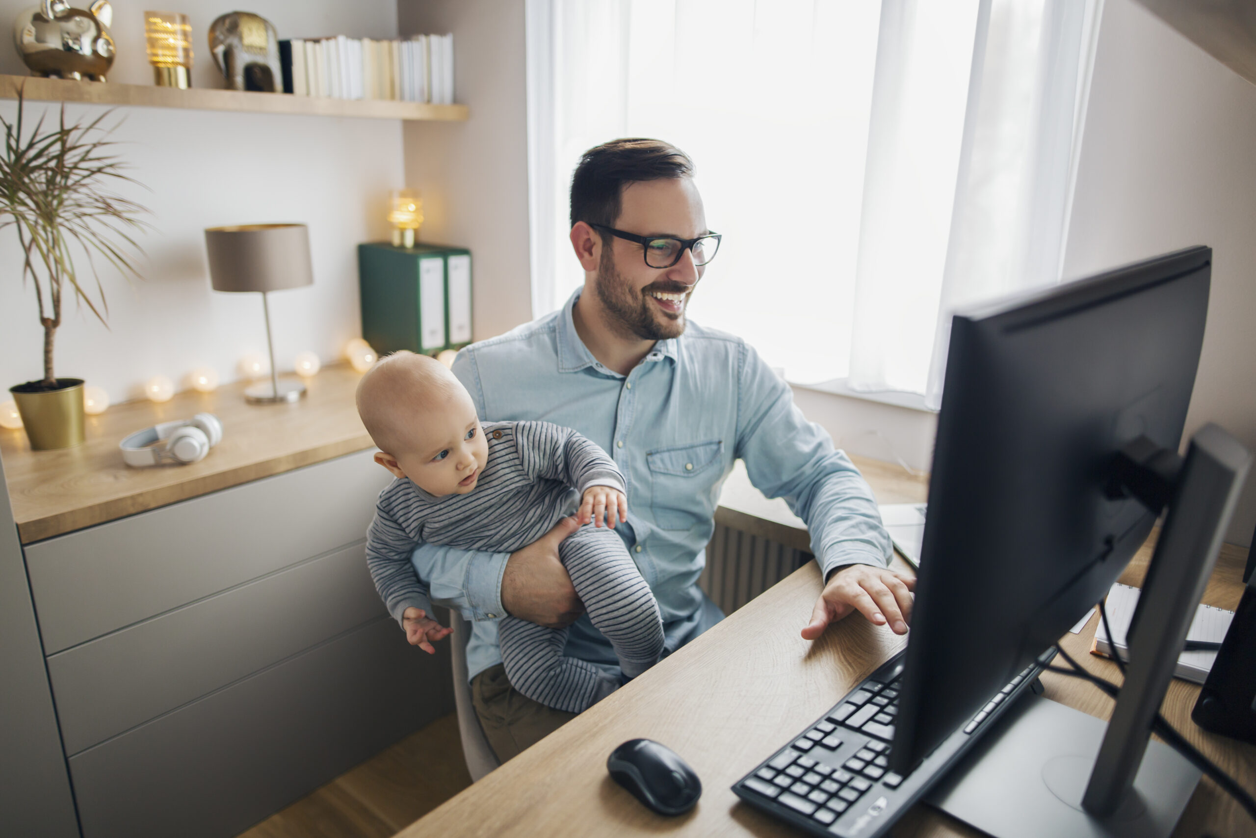 Young smiling father works from home typing on his computer while holds his son while they both watch screen.